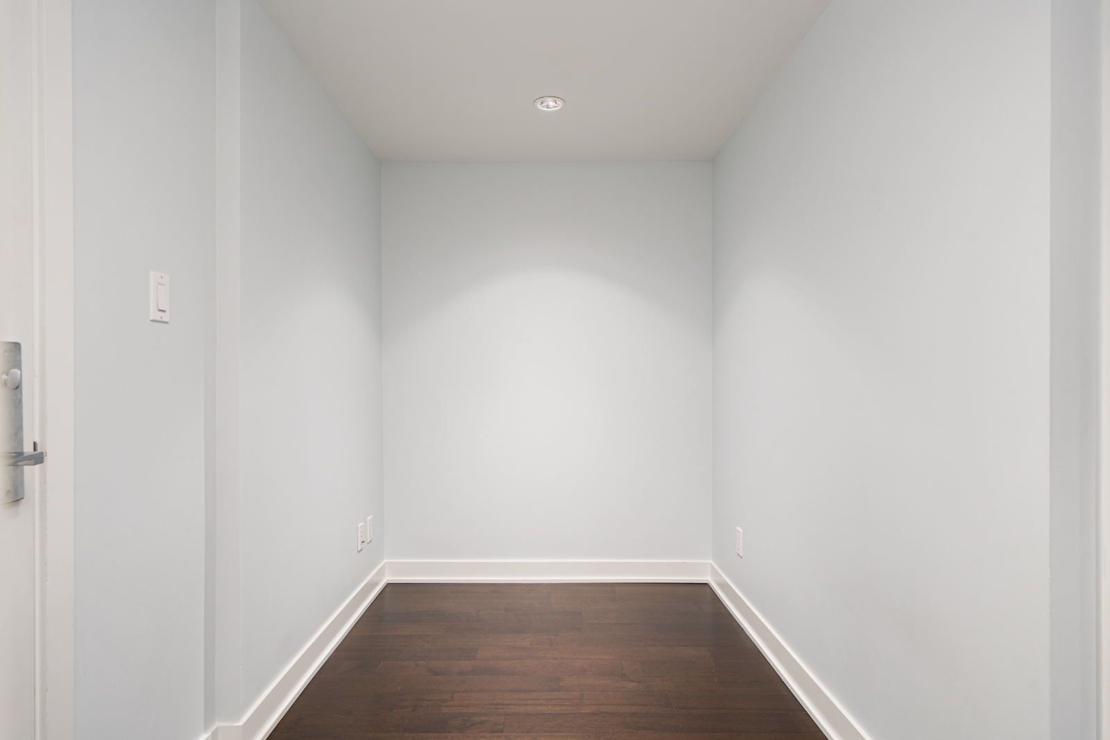 A small, empty room with white walls, dark wooden floor, and a single ceiling light.