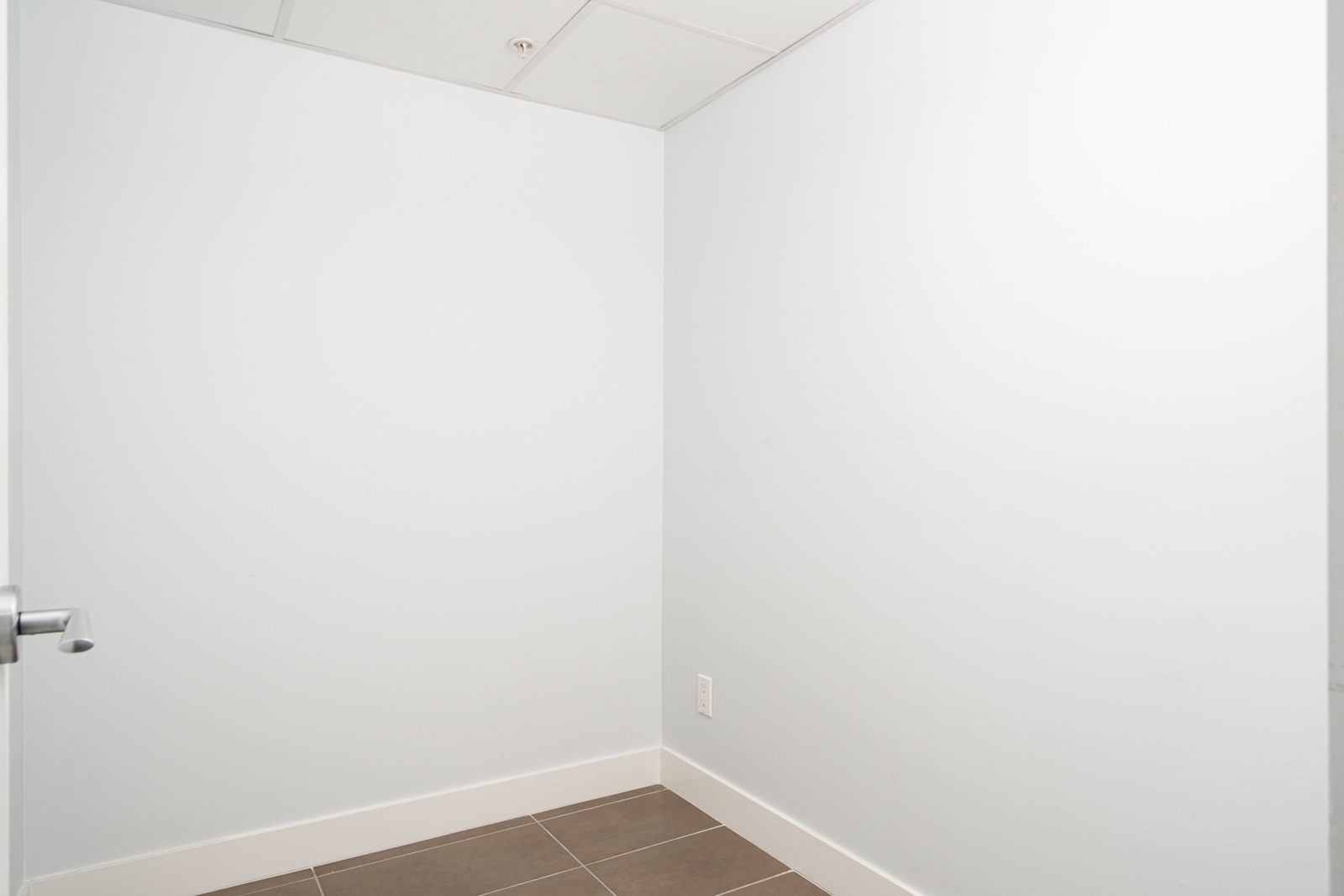 Empty corner of a room with white walls, tiled floor, and a silver door handle on the left.