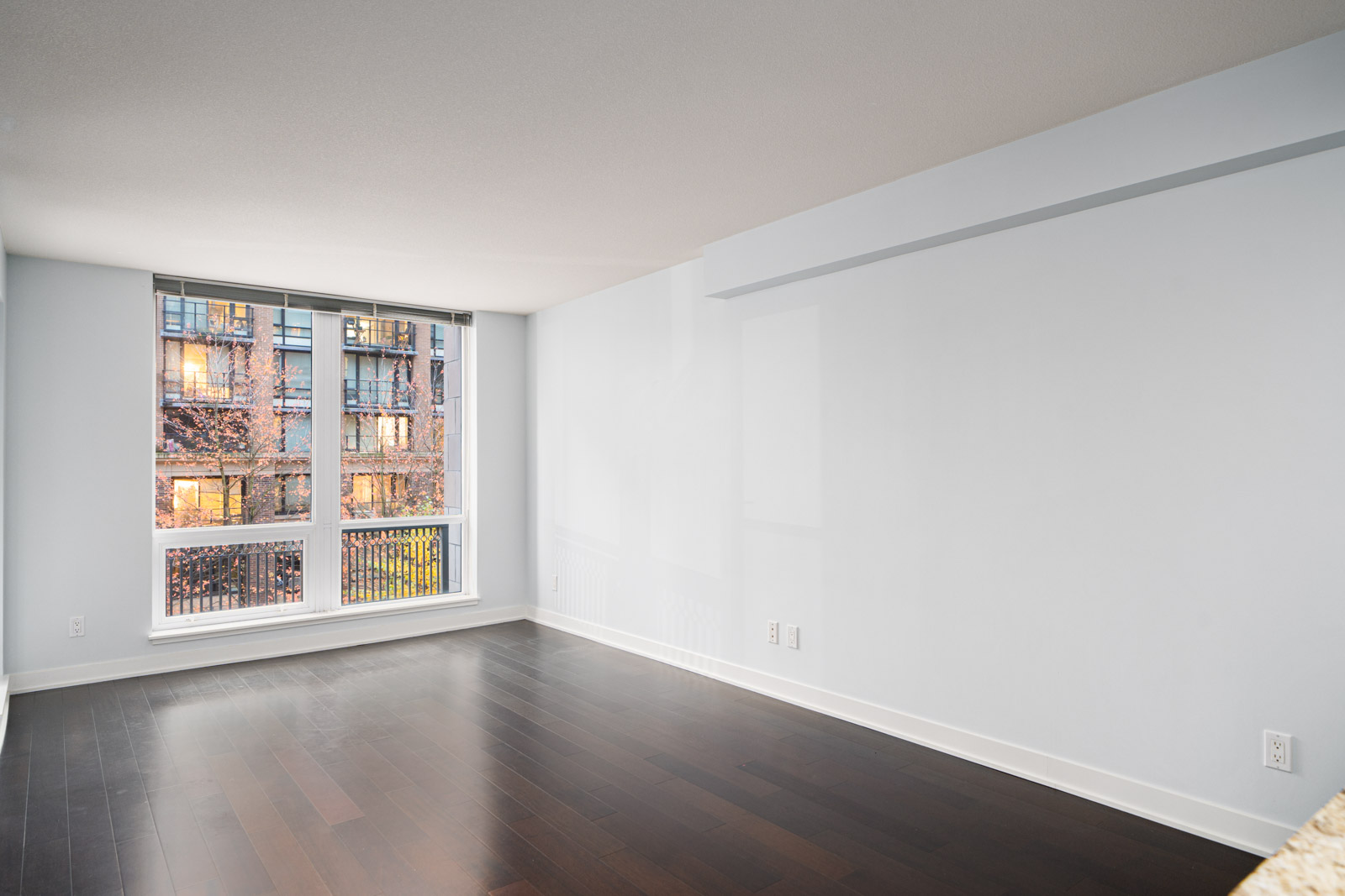 Empty room with dark wooden floor, white walls, and large windows overlooking a building exterior.