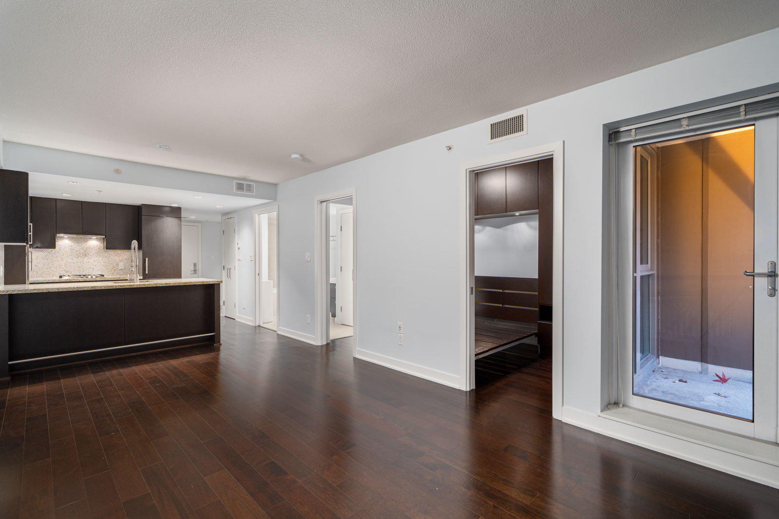 Modern apartment interior with dark wood flooring, an open kitchen with dark cabinets, and a sliding glass door leading to a small outdoor space. Three doors lead to additional rooms.