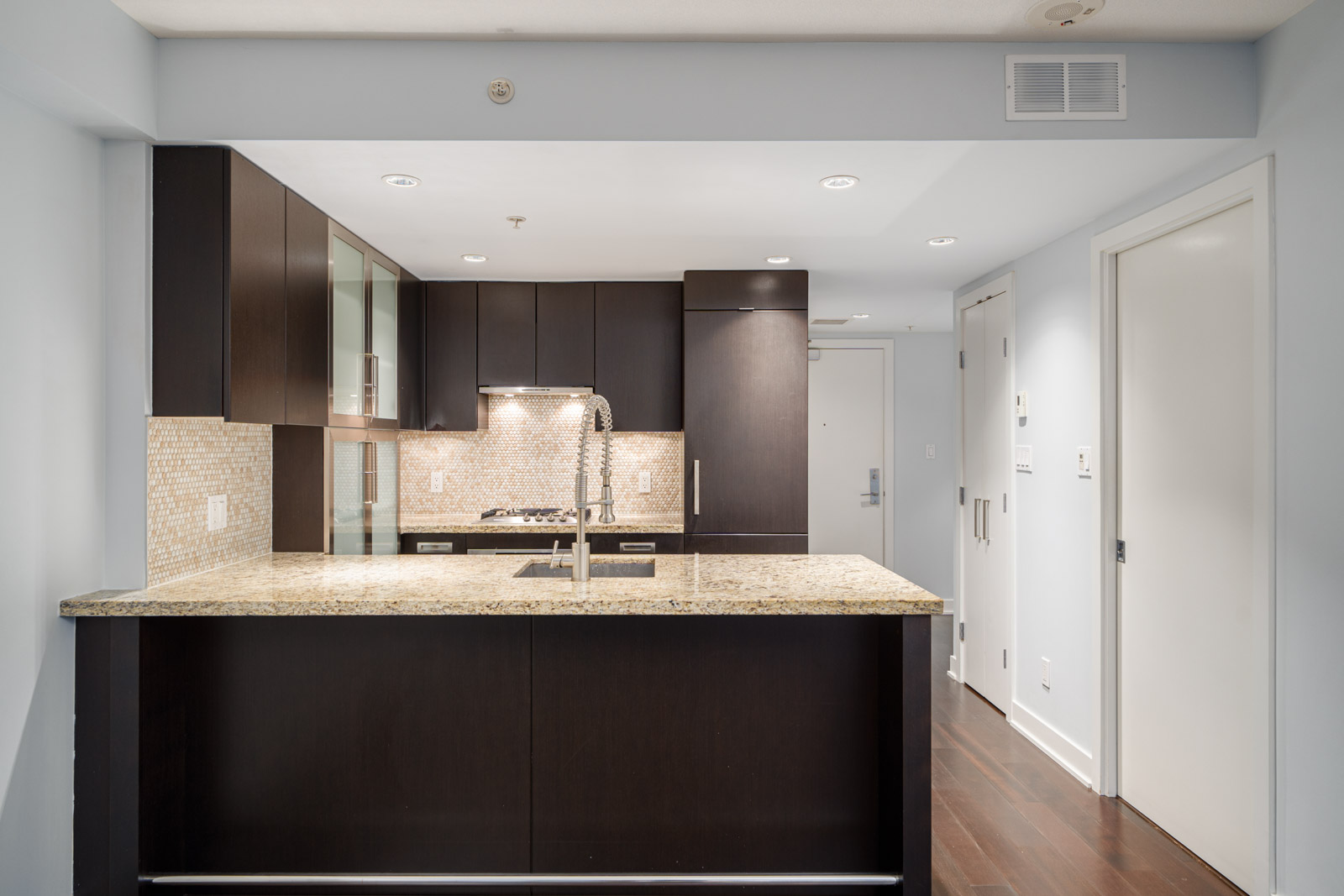 A modern kitchen with dark cabinetry, a light granite countertop, and a stainless steel sink. The backsplash is tiled, and recessed lighting is installed in the ceiling.