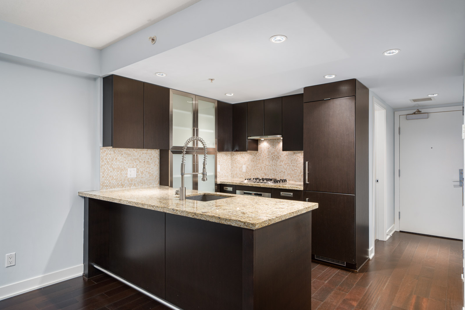 Modern kitchen with dark wood cabinets, granite countertops, a central island with a double sink, and a tile backsplash. Ceiling lights illuminate the space.