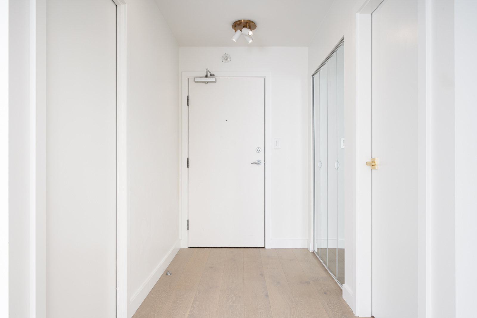 A white hallway with a wooden floor, featuring a closed door at the end, a mirrored sliding closet door on the right, and a wall-mounted light fixture on the ceiling.