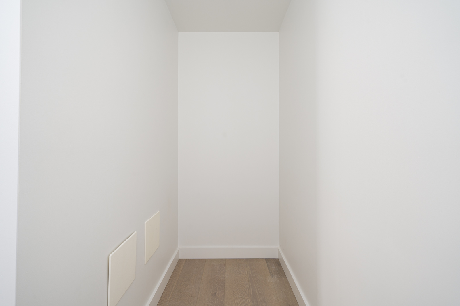 Narrow empty white corridor with wooden floor and two rectangular panels on the left wall.