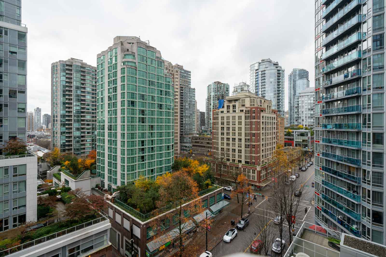 View of a cityscape with tall modern buildings, roads, and trees with autumn foliage. Cloudy sky overhead.