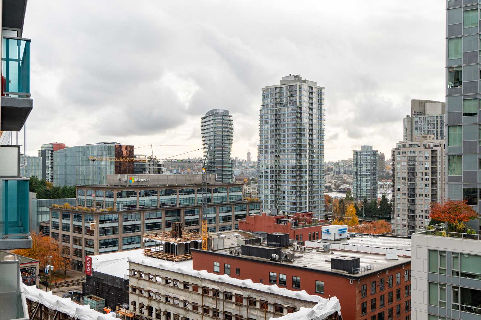 Cityscape with modern high-rise buildings, a construction crane, and overcast sky. Trees with autumn colors are visible among the structures.