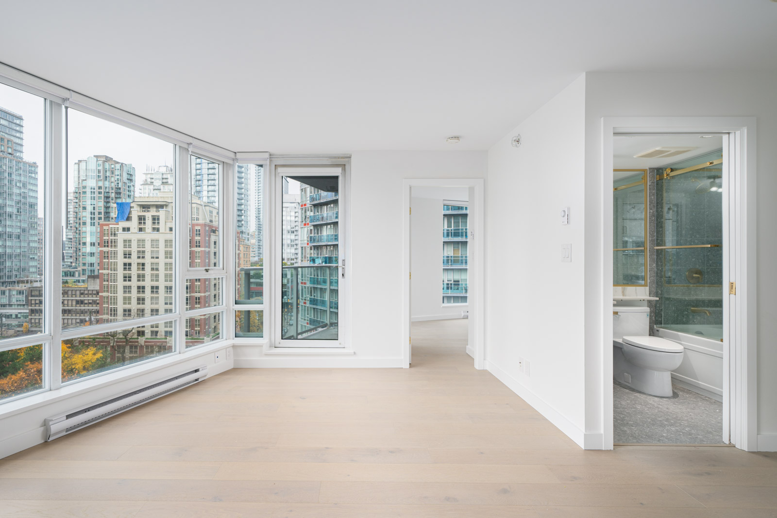 Bright apartment with large windows, light wood floors, and a view of city buildings. Includes a white-walled bathroom with a shower and toilet.