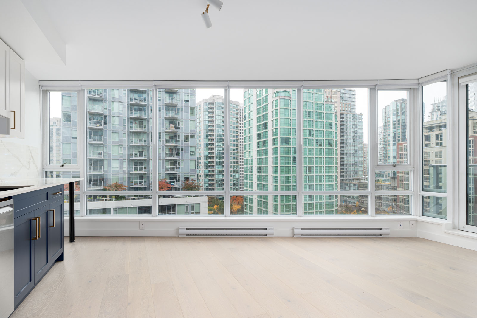 Bright room with large windows showcasing a cityscape of high-rise buildings. Light hardwood floors and modern kitchen cabinetry are visible.
