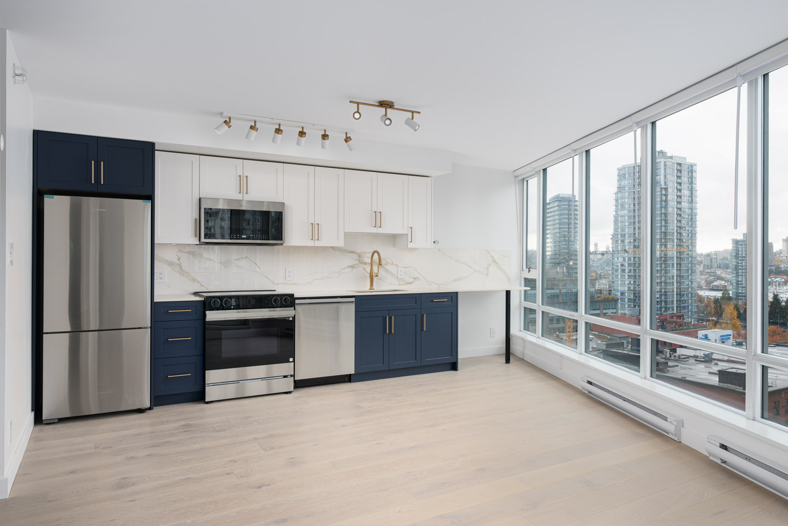 Modern kitchen with navy blue and white cabinets, stainless steel appliances, and a marble backsplash. Large windows offer a city view with ample natural light.