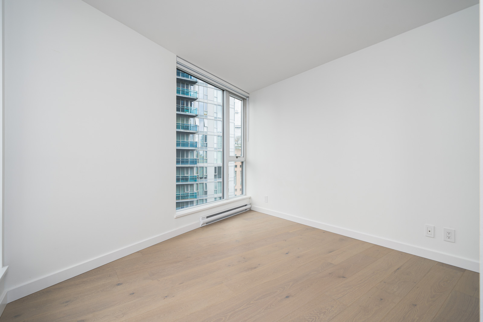 Bright, empty room with light wood flooring, white walls, and a large window showing modern buildings outside.