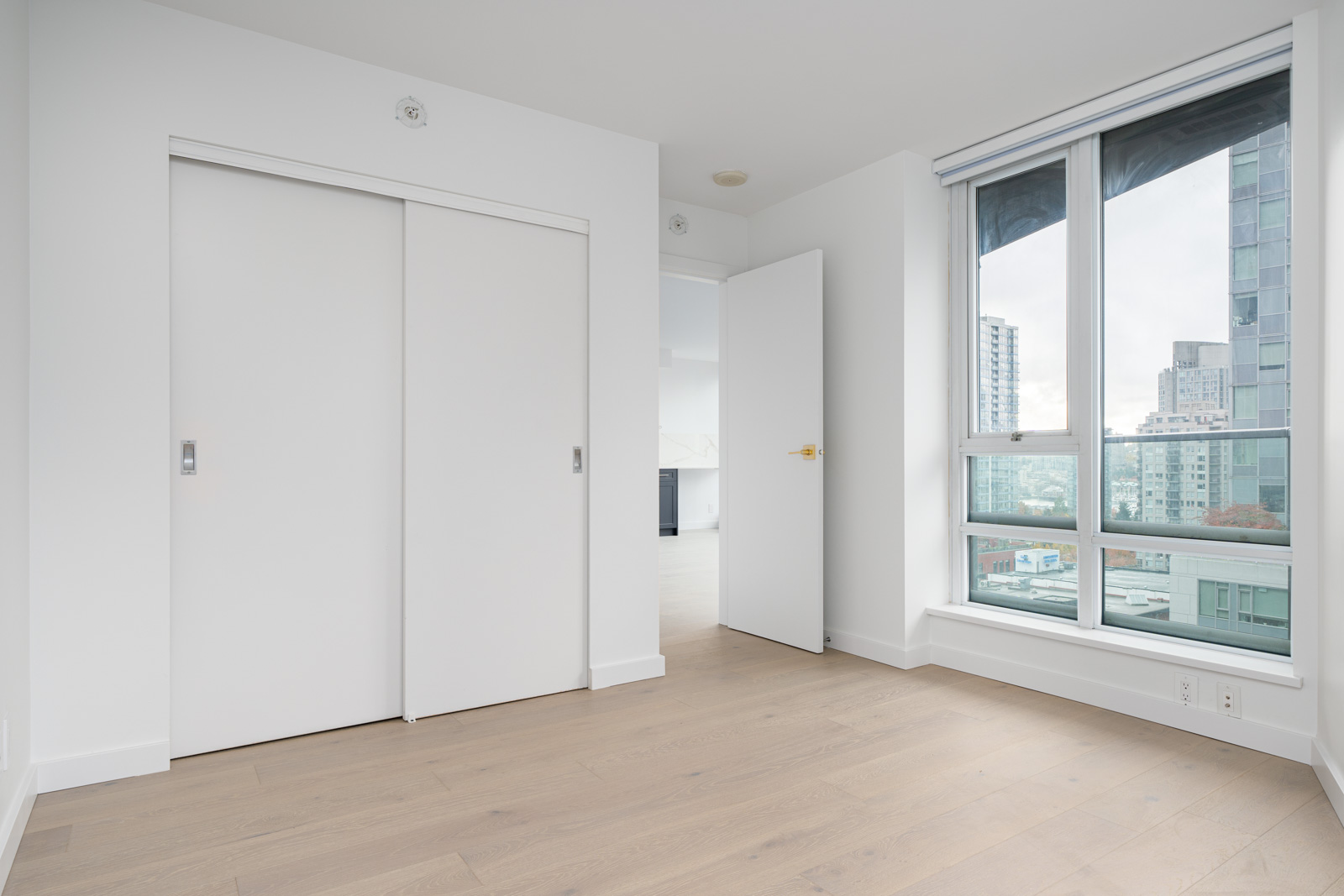 Empty room with light wood flooring, white walls, sliding closet doors, an open door, and a large window with a city view.
