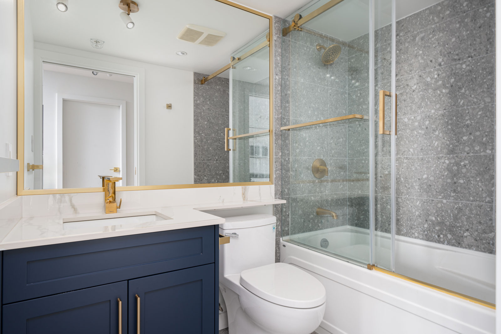 Modern bathroom with a navy blue vanity, white countertop, and gold fixtures. Glass-enclosed bathtub with gray tile wall and sliding door.