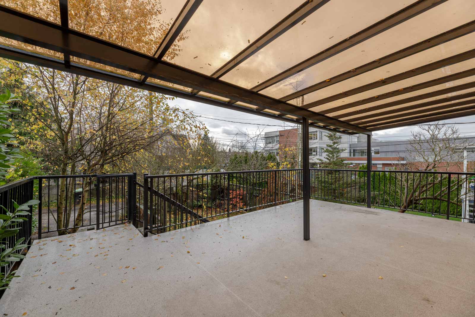 Covered balcony with a transparent roof and metal railings, overlooking a tree-lined street. Nearby foliage is visible under an overcast sky.