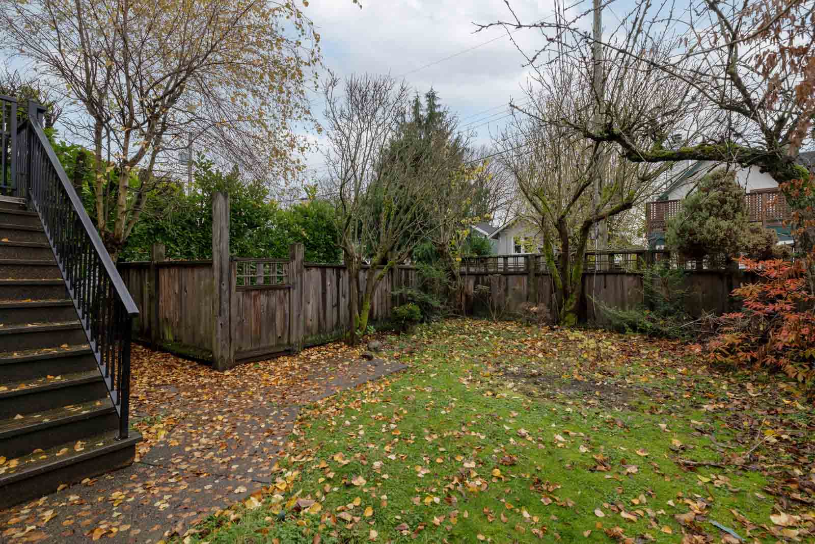 Backyard with a wooden fence, leaf-covered grass, trees, and a staircase on the left. The sky is overcast.