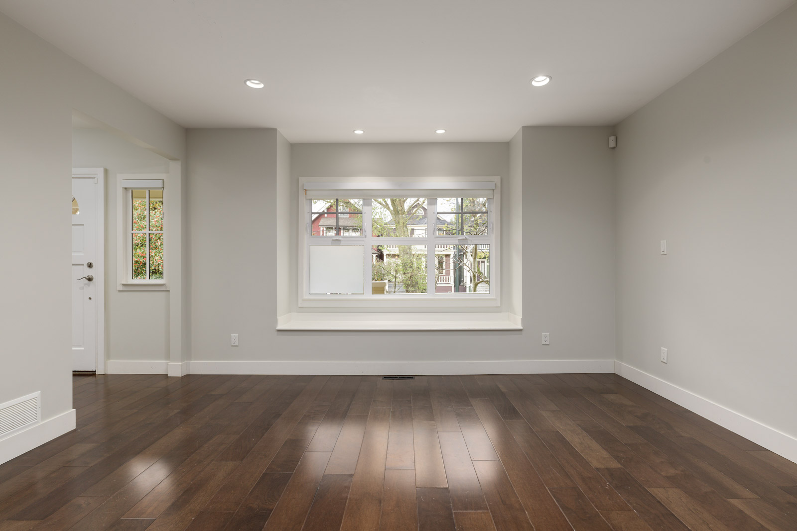An empty room with beige walls, recessed lighting, and dark wooden floors. A large window with a view of trees is on the far wall.