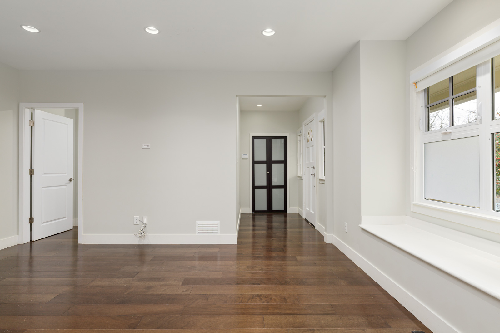 Empty room with hardwood floor, white walls, and recessed lighting. Includes a closed door, a window with a sill, and a dark framed doorway.