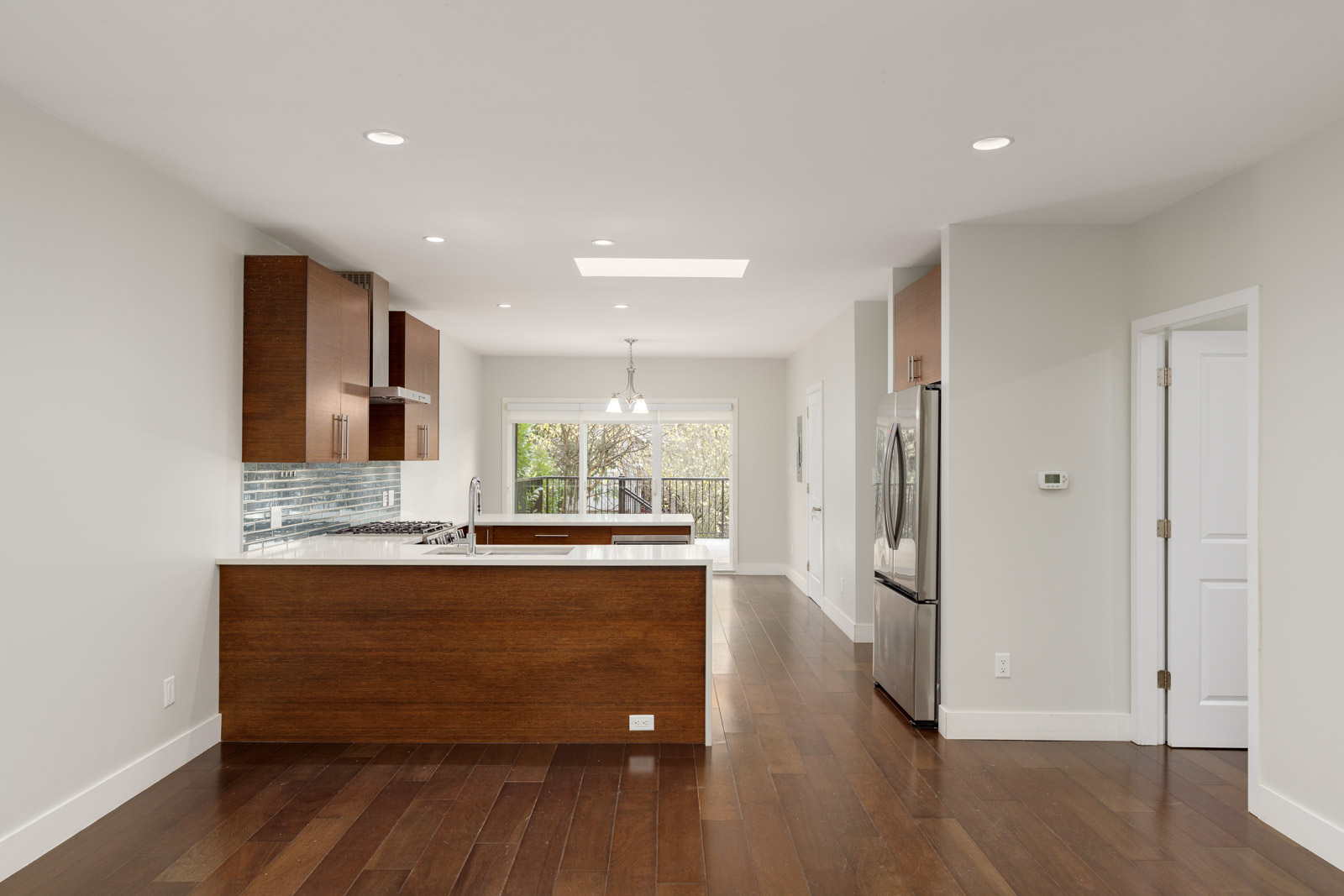 Modern kitchen with wooden cabinets, stainless steel appliances, and hardwood floors. A glass door at the back opens to a patio, with recessed lighting and neutral walls completing the decor.
