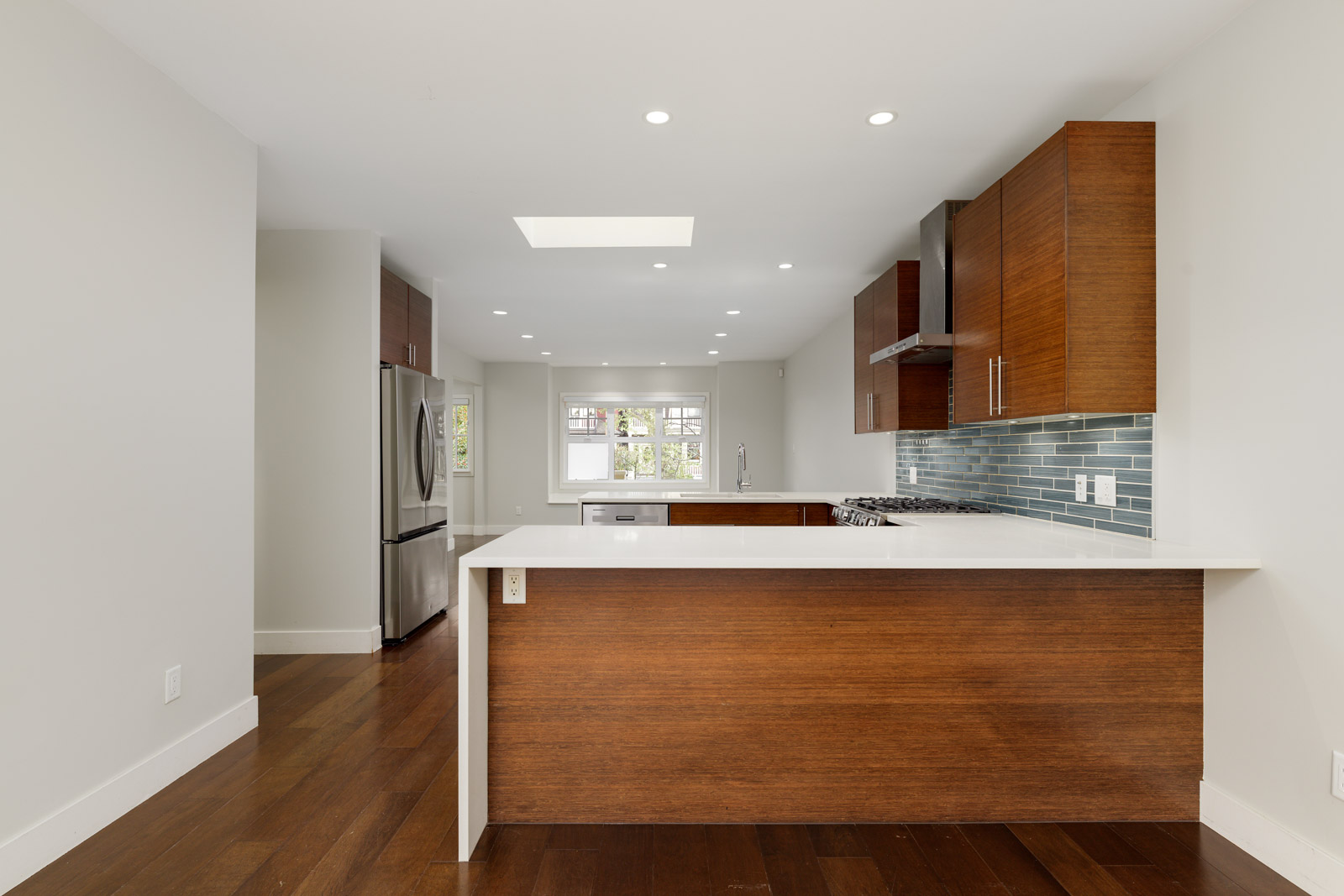 Modern kitchen with wooden cabinetry, a white countertop, stainless steel appliances, and a blue tiled backsplash. The room is bright with recessed lighting and an open layout.