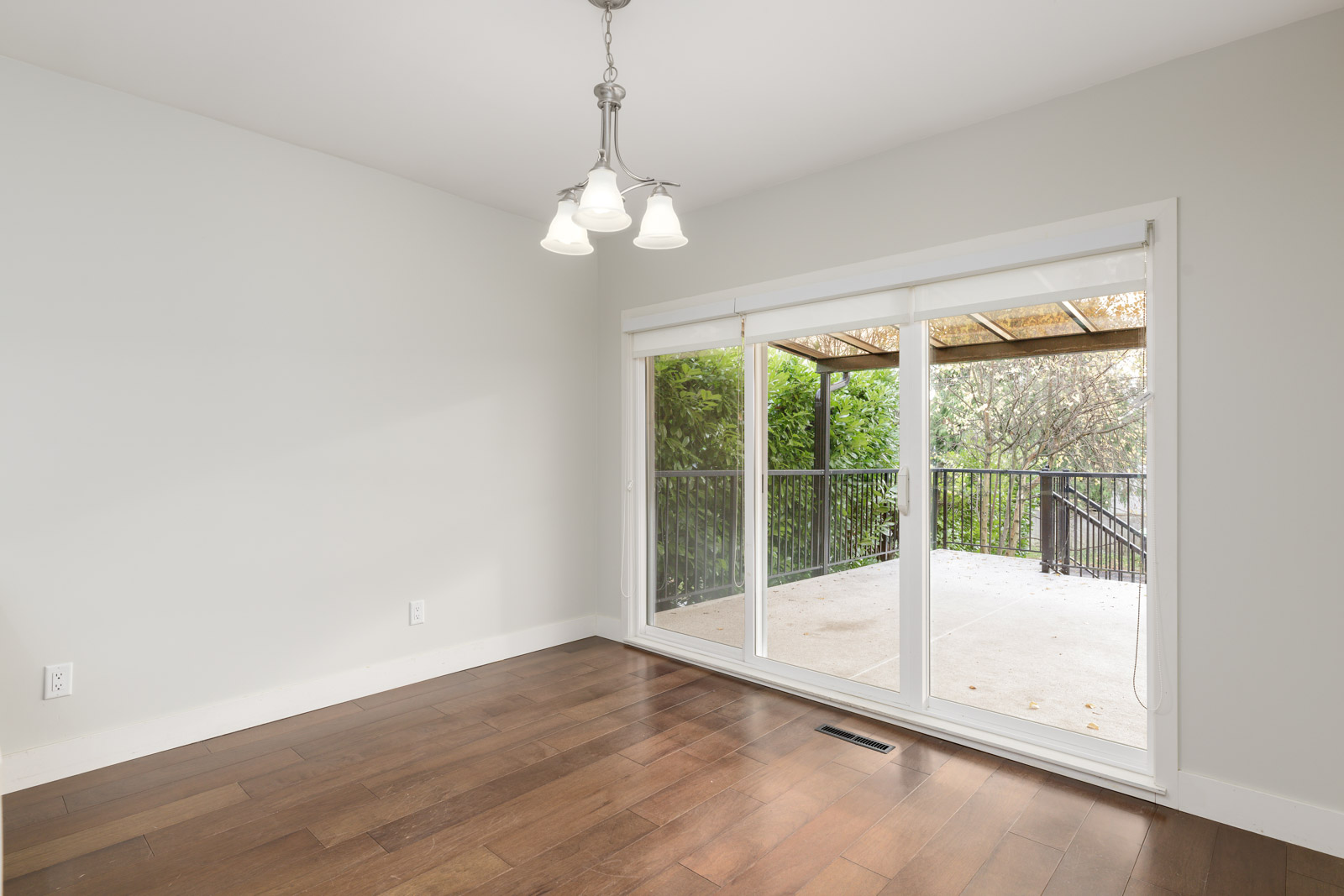 A bright, empty room with hardwood floors, a pendant light, and large sliding glass doors leading to an outdoor patio with a railing.