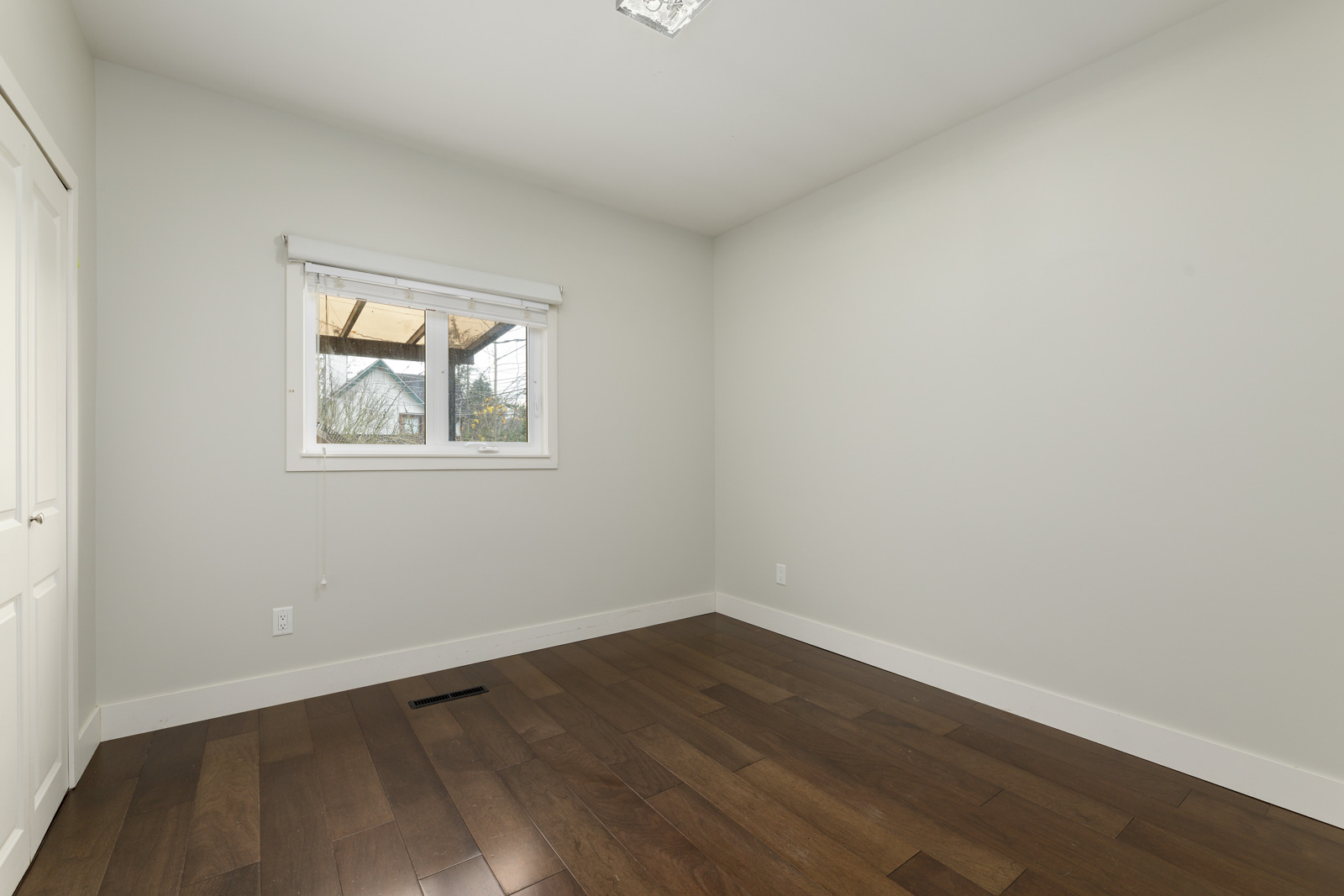 Empty room with light gray walls, a window with a blind, and dark wood flooring.