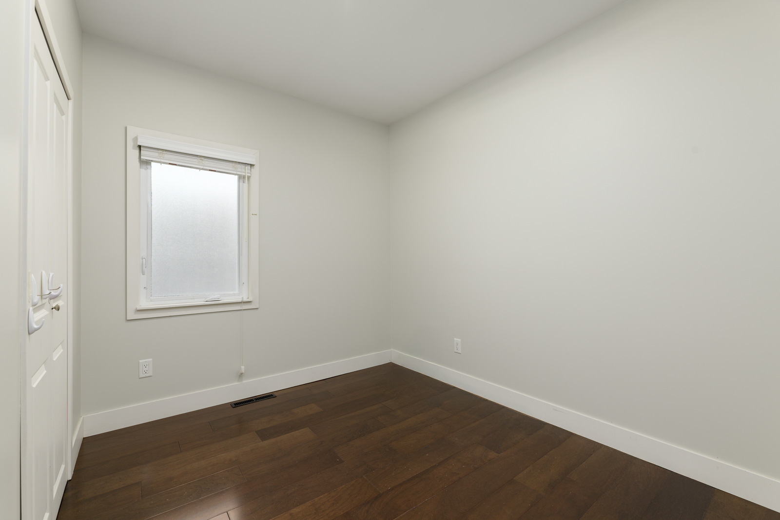 Empty room with cream walls, a small window with white trim, and dark hardwood flooring. A white closet door is partially visible on the left.