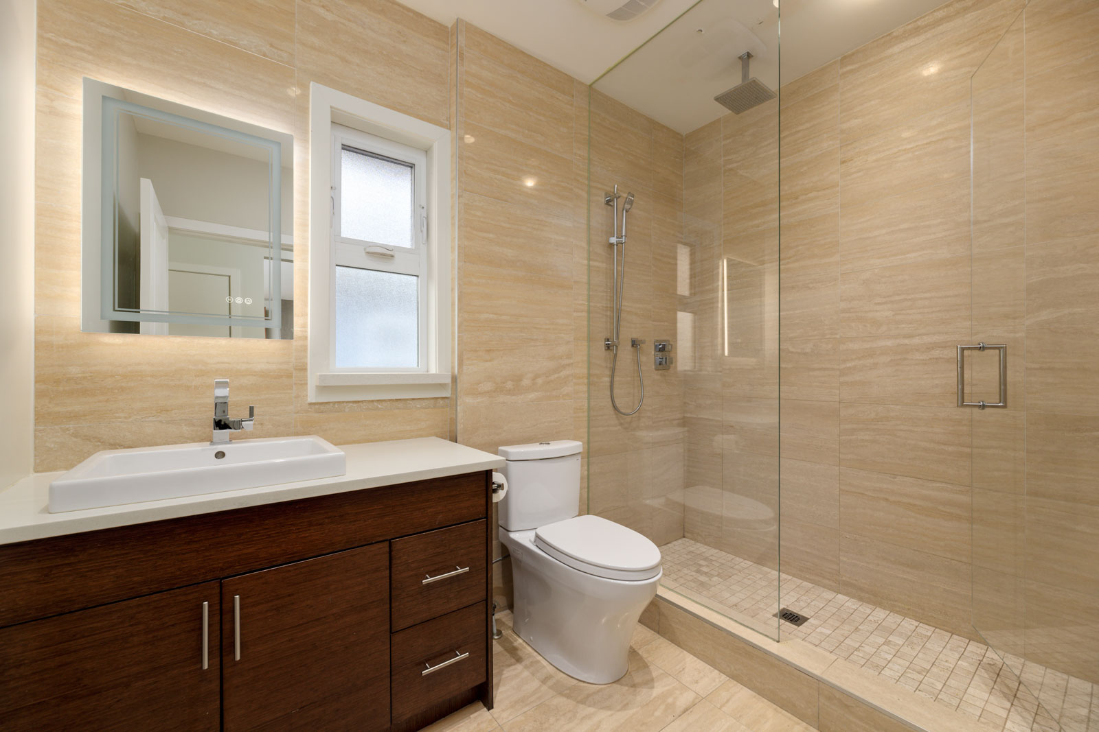 Modern bathroom with beige tiles, a glass-enclosed shower, a white toilet, a dark wood vanity with a white sink, and a rectangular mirror.