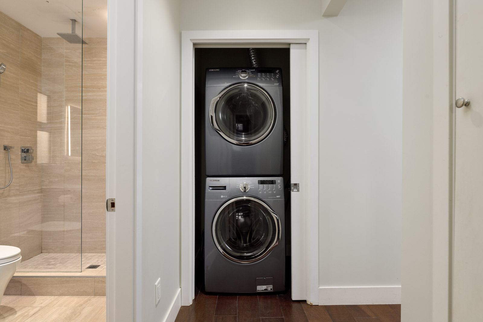 Stacked washer and dryer in a narrow closet next to a bathroom with a glass shower and wood flooring.