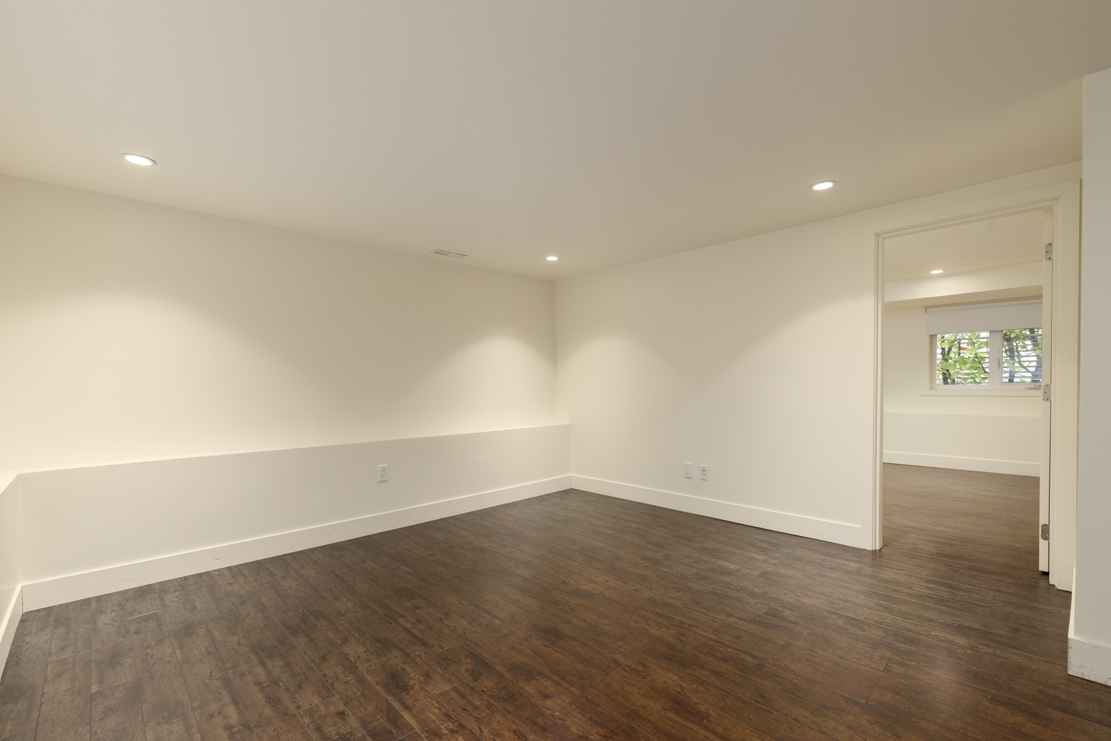 A clean, empty room with light beige walls, dark wood flooring, and recessed lighting. A doorway leads to another room.