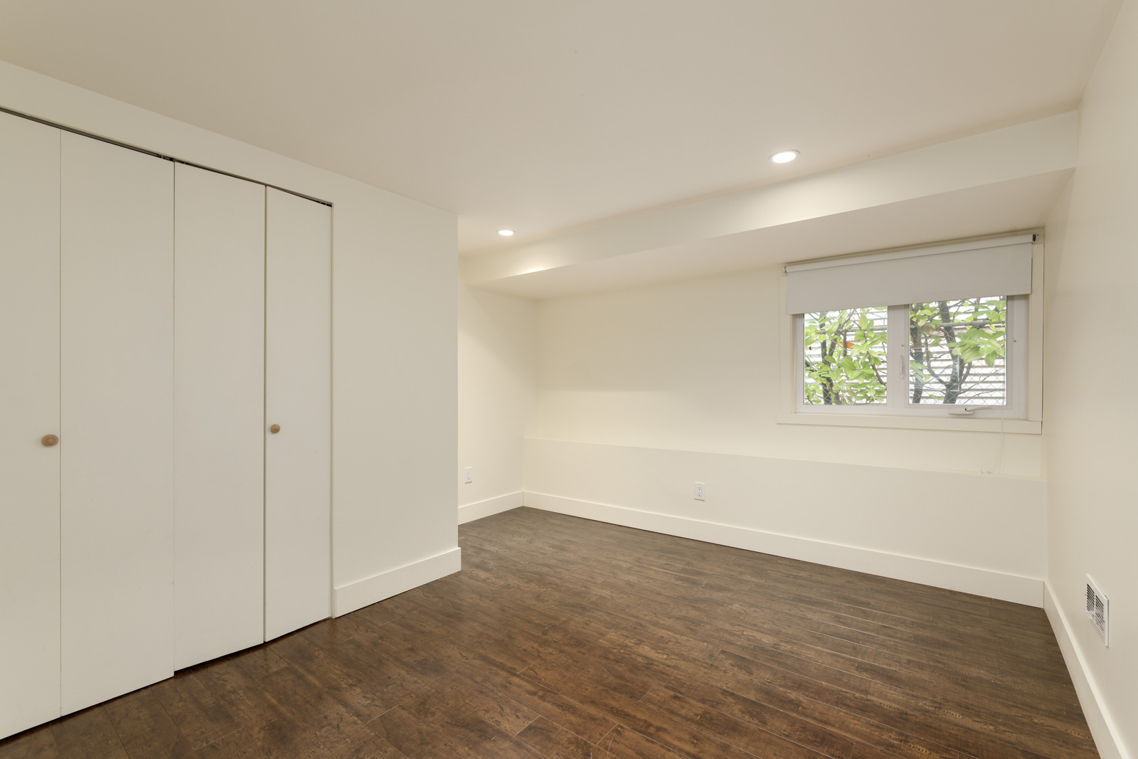 An empty room with white walls, a small window with blinds, a wooden floor, and a closed closet with sliding doors.