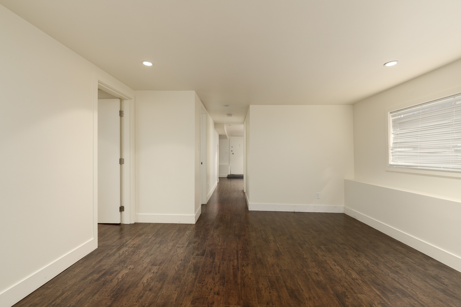 A narrow hallway with white walls, dark wooden floor, and a window with blinds on the right. There is a doorway on the left and recessed lighting on the ceiling.