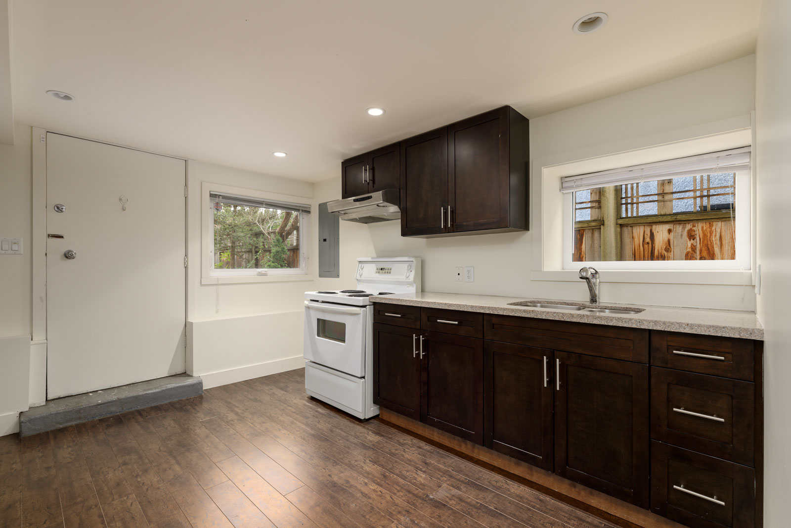 A small kitchen with dark wood cabinets, white electric stove, and a sink. Two windows provide natural light. Doorway and wood flooring are visible.