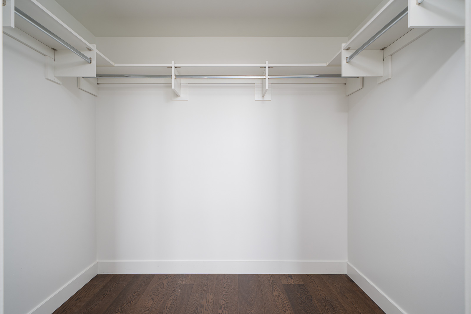 Empty walk-in closet with white walls, shelving, and hanging rods, featuring a dark wooden floor.