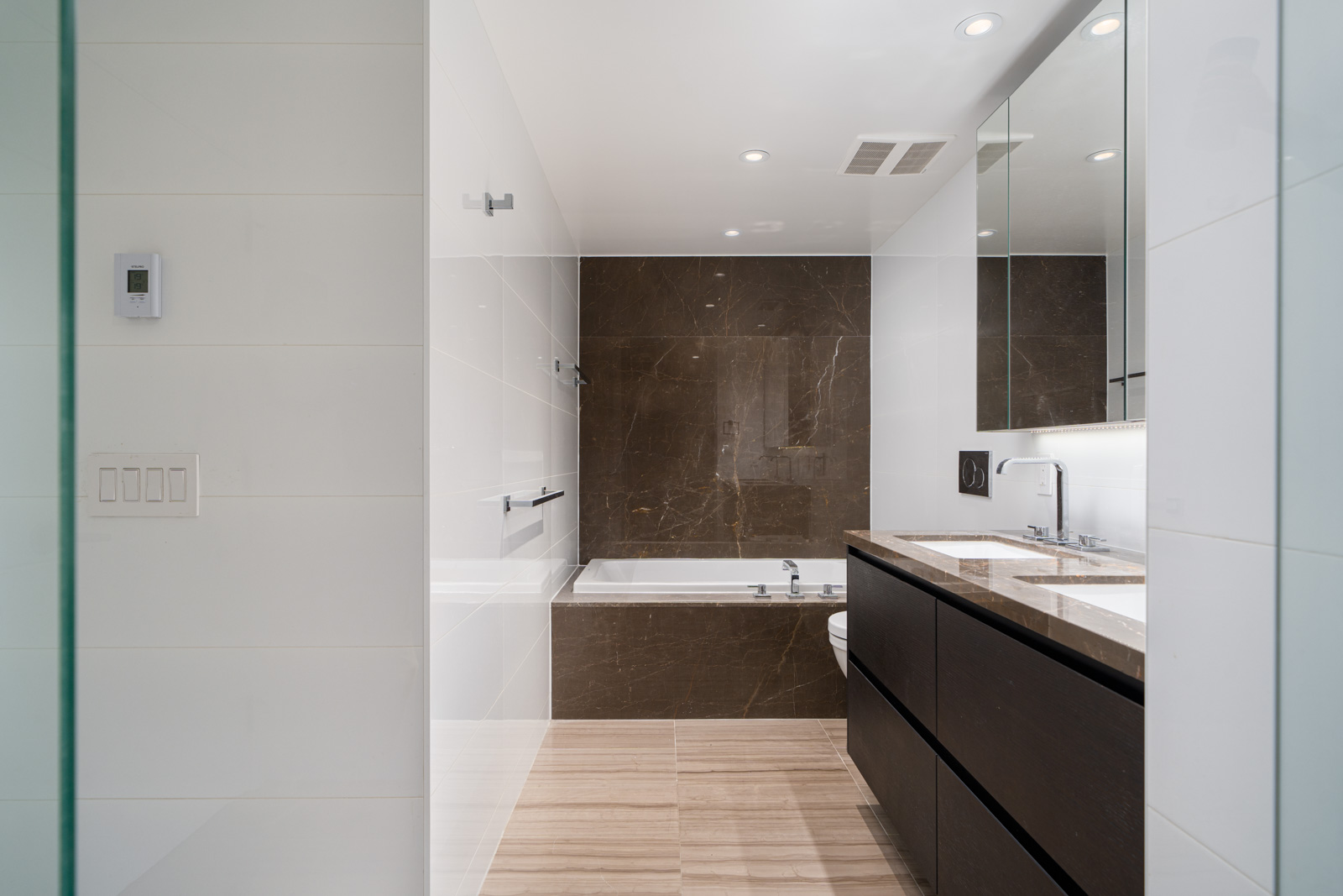 Modern bathroom with a bathtub, dark wood cabinets, marble backsplash, double sinks, and large mirrors. White and beige color scheme with recessed lighting.