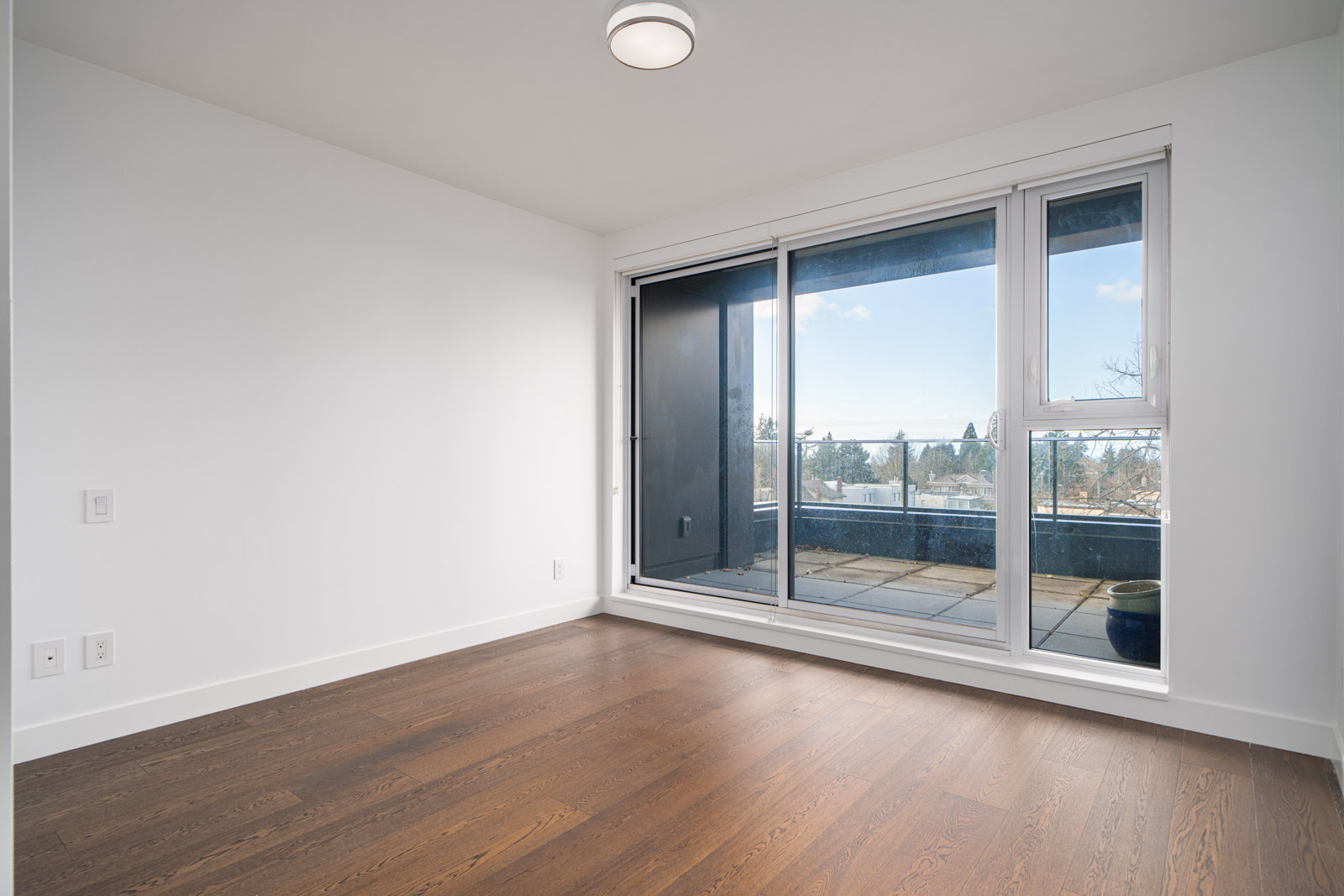Empty room with wooden floors, white walls, and a large glass sliding door leading to a balcony.