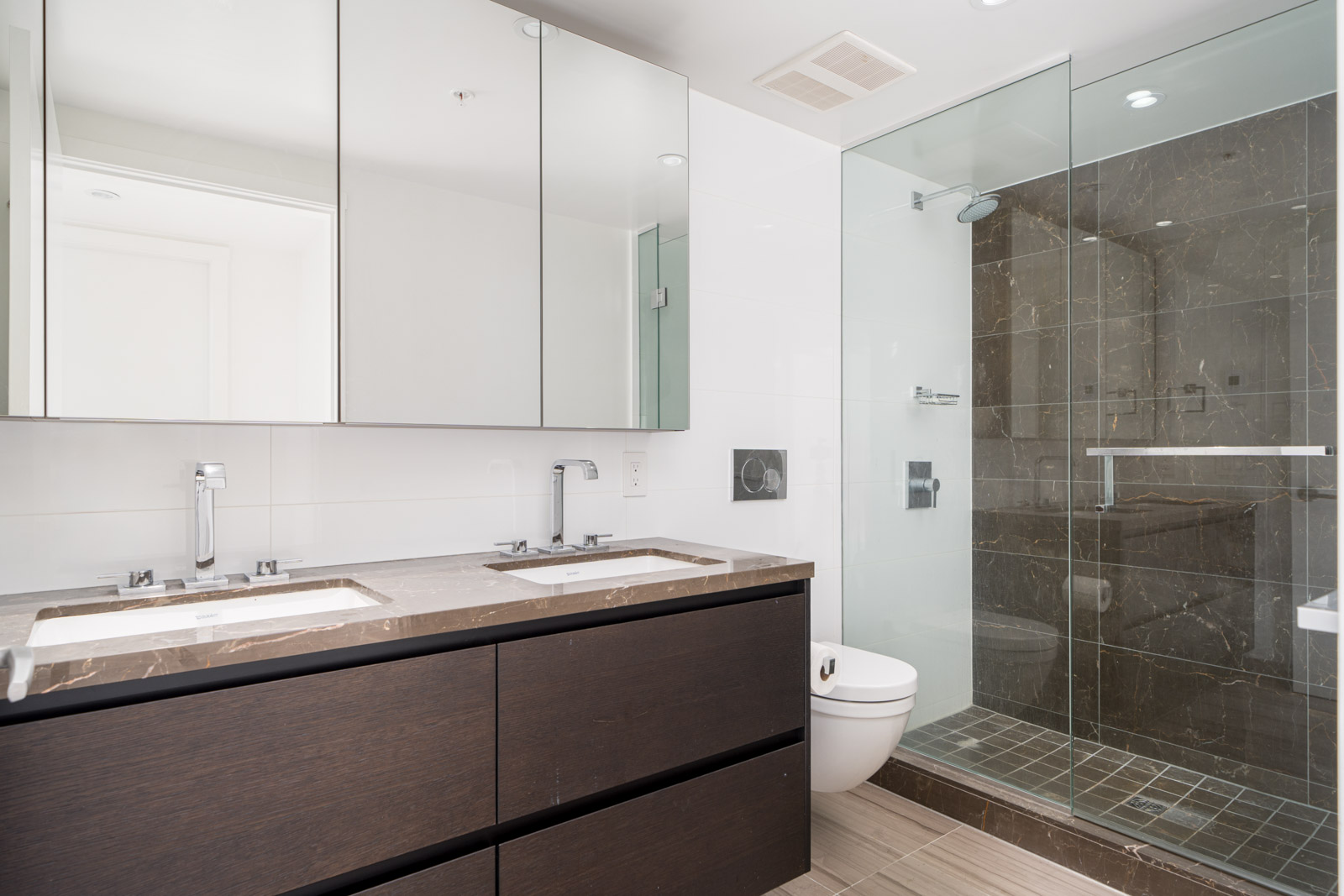 Modern bathroom with dark wood double vanity, large mirrors, glass-enclosed shower with dark tiles, and wall-mounted toilet.