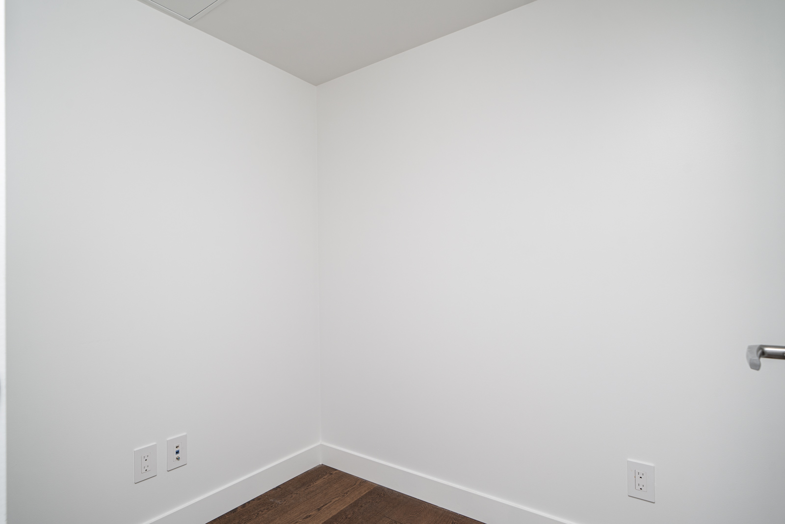 Small, empty room with white walls, a wooden floor, and electrical outlets.