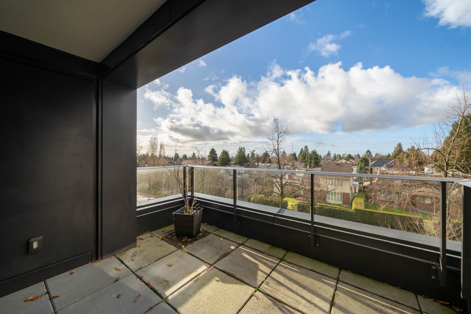 A spacious balcony with glass railings offers a view of a suburban neighborhood with trees and houses under a cloudy blue sky.