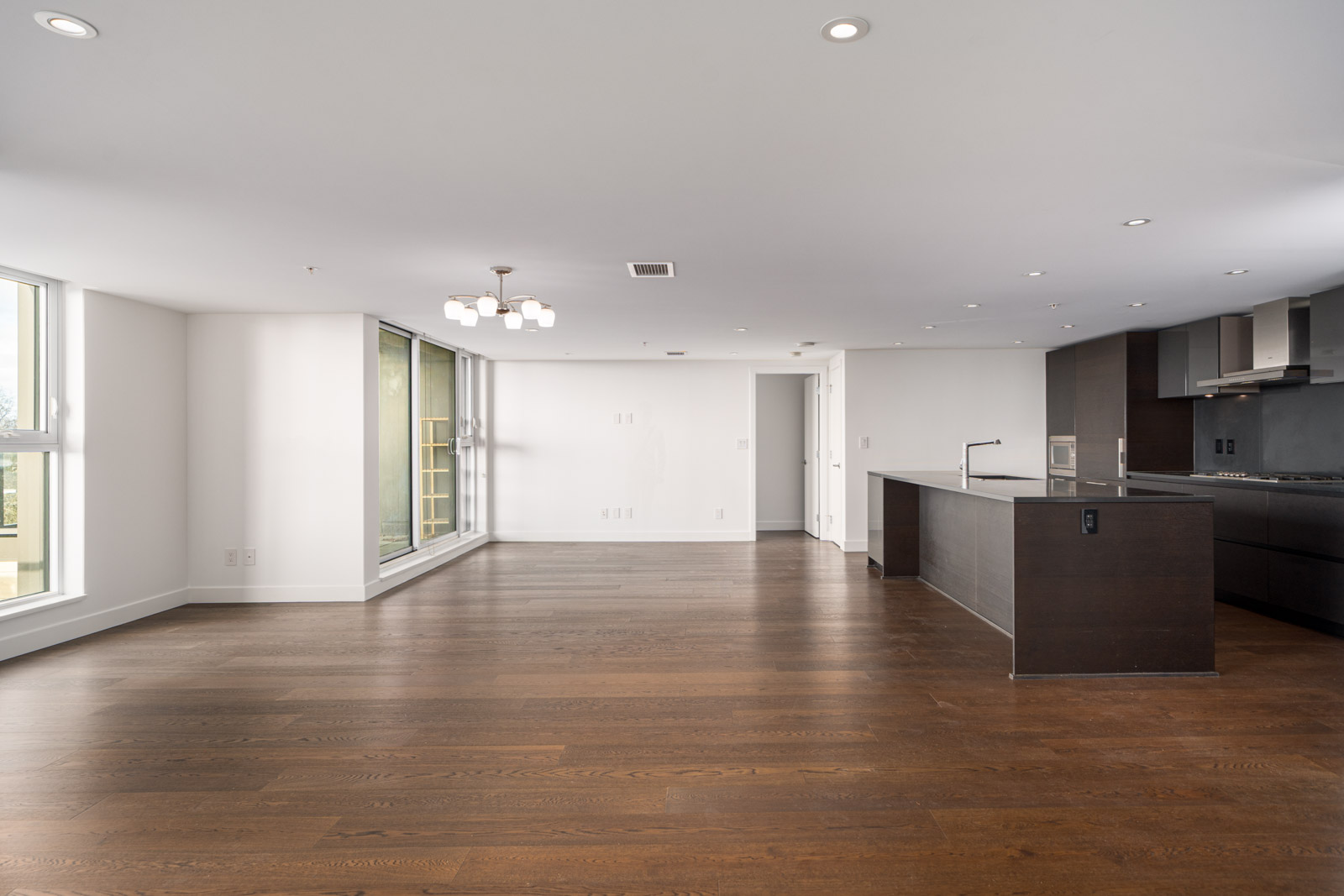 Spacious modern apartment interior with hardwood flooring, large windows, minimalist lighting fixture, and an open kitchen with dark cabinets and an island.