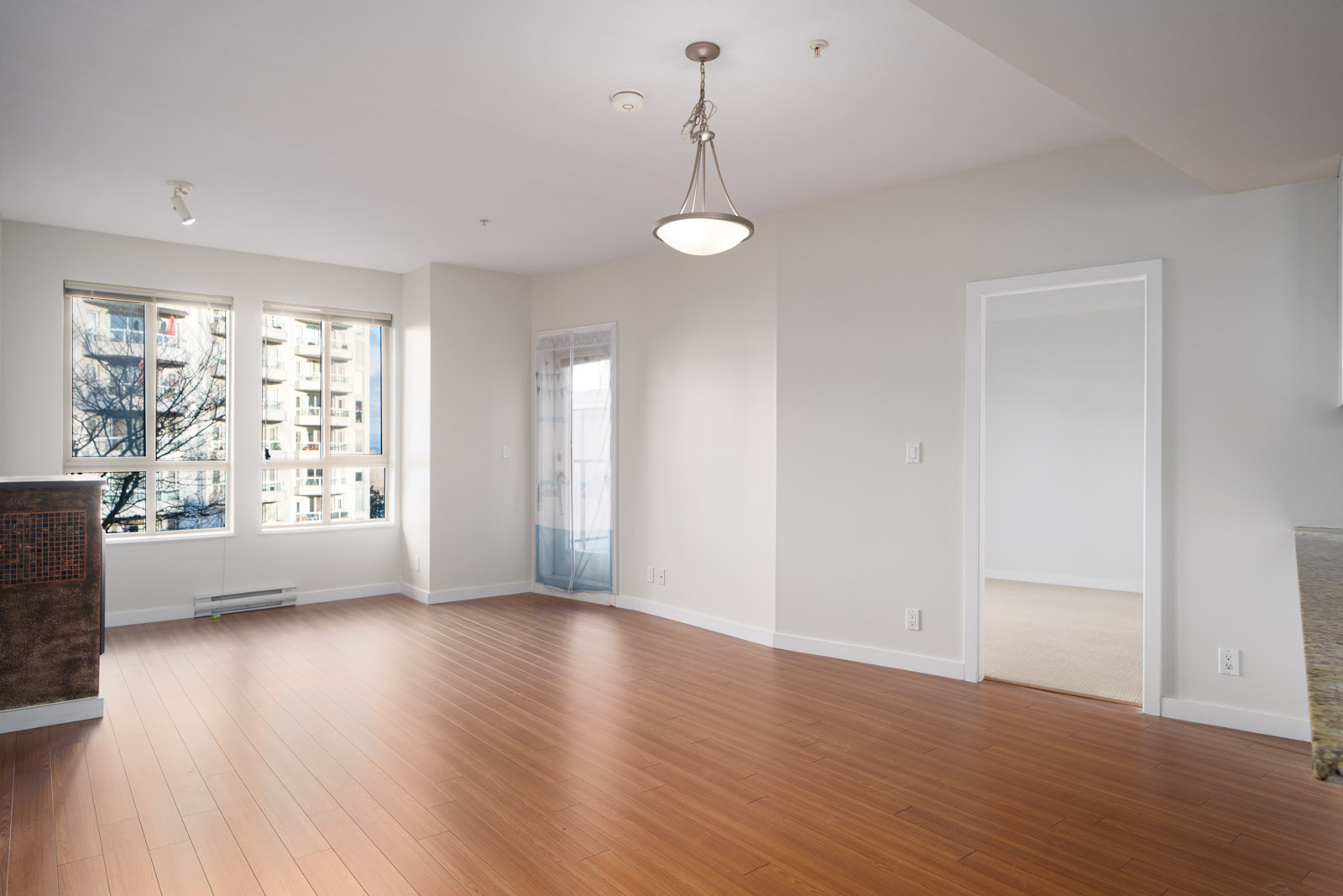 A spacious, empty room with hardwood floors, large windows, neutral walls, and a hanging light fixture. Another room is visible through an open doorway.