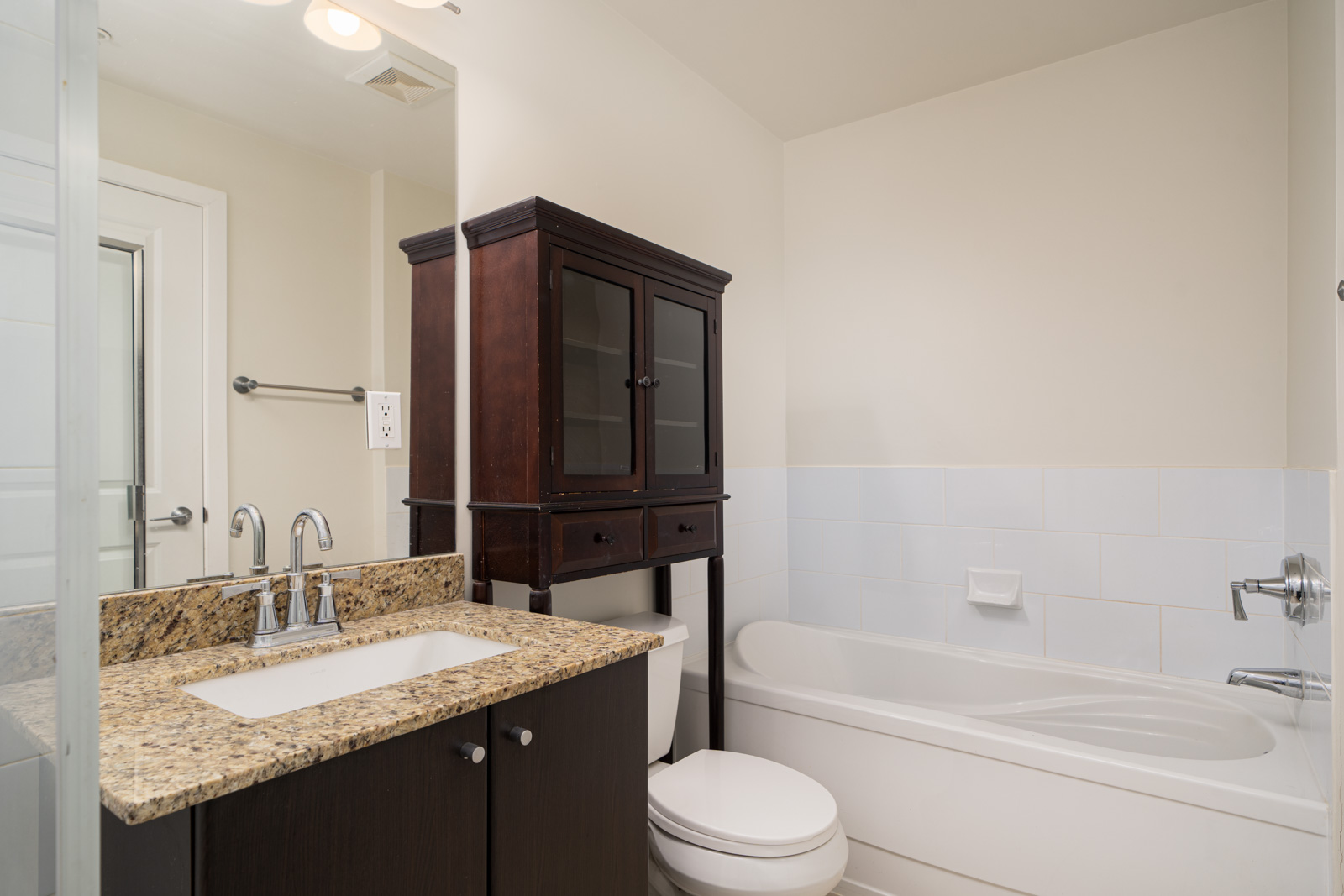 A bathroom with a granite countertop sink, a wooden cabinet, a toilet, and a bathtub with white tiles.
