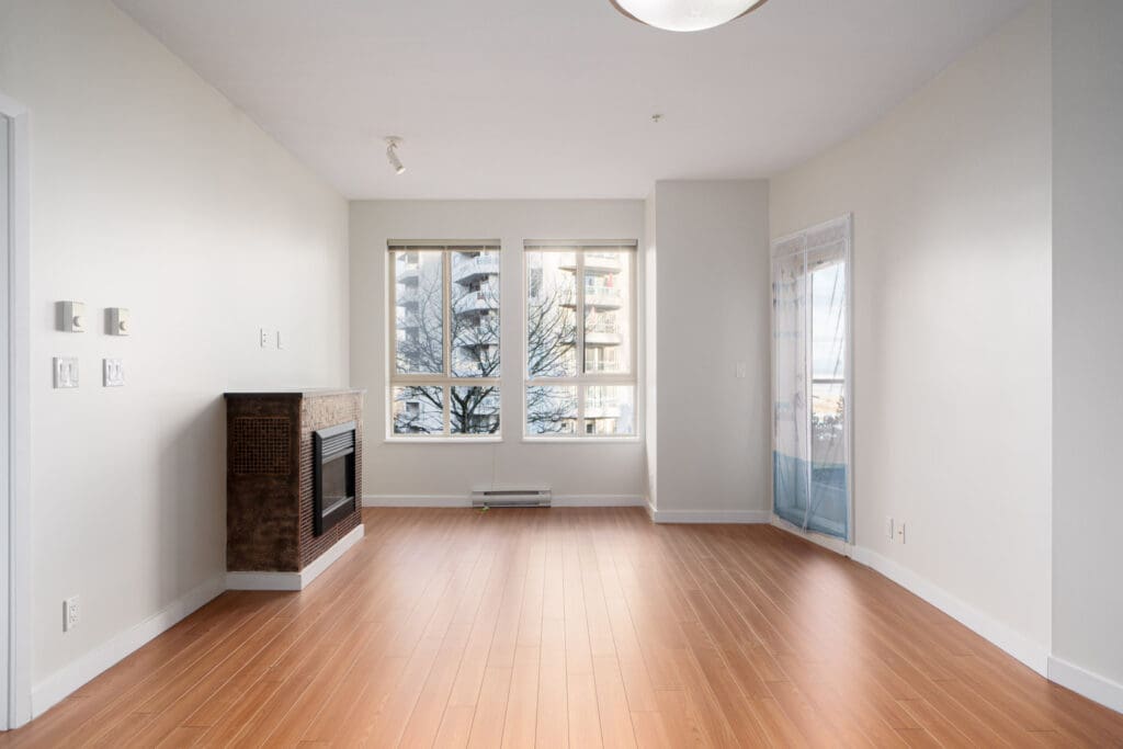 An empty, bright living room with wood flooring, large windows, a fireplace, and a glass door leading to a balcony.