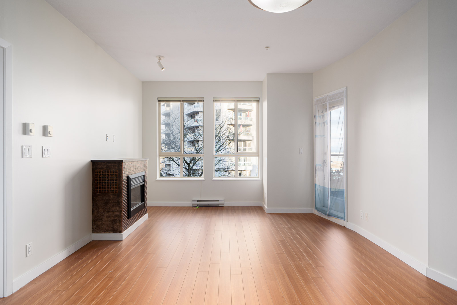 An empty, bright living room with wood flooring, large windows, a fireplace, and a glass door leading to a balcony.