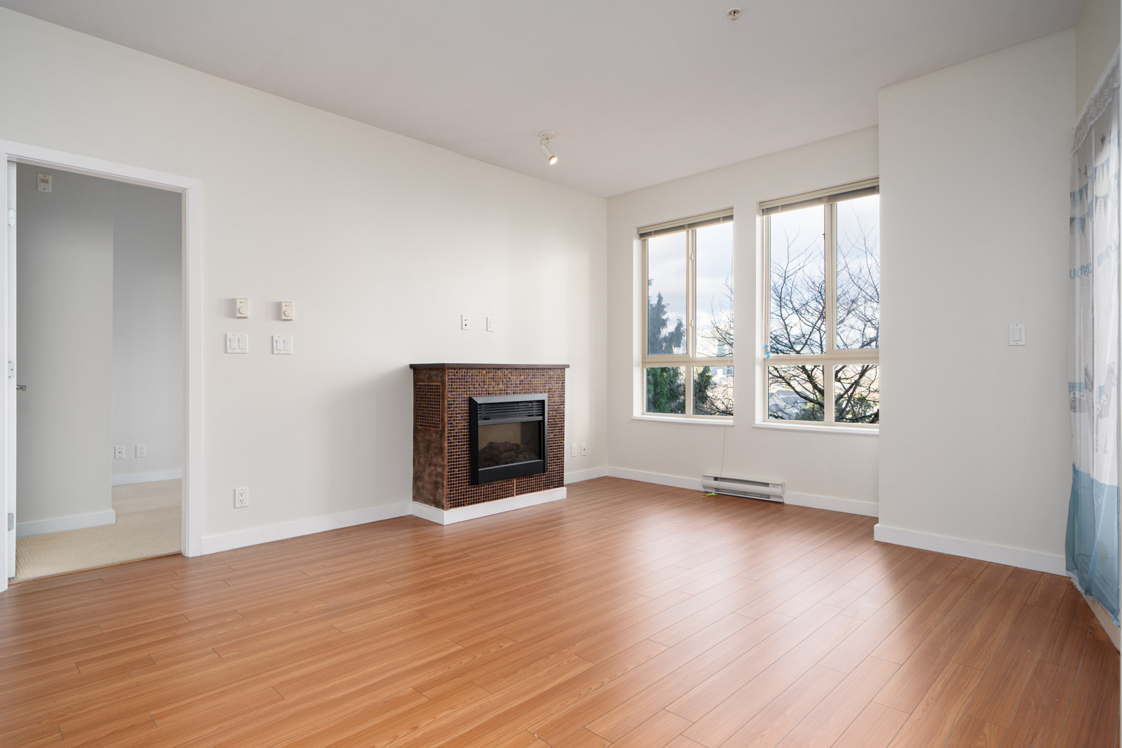 Empty living room with wood flooring, a fireplace, and large windows. White walls, natural light, and an open doorway to an adjacent room.
