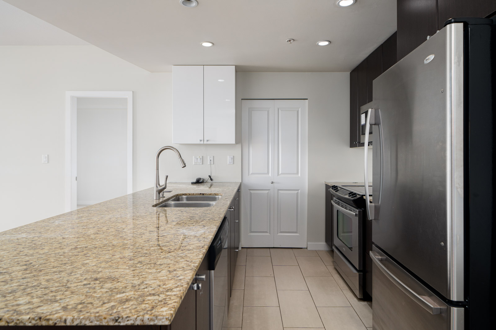 Modern kitchen with granite countertop, stainless steel appliances including stove and refrigerator, dark cabinets, and beige tiled floor.