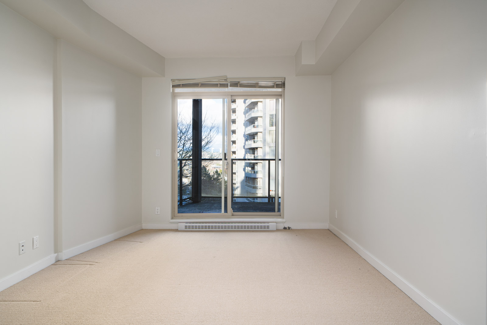 Empty room with beige carpet, white walls, and a large window leading to a balcony. A high-rise building is visible outside.
