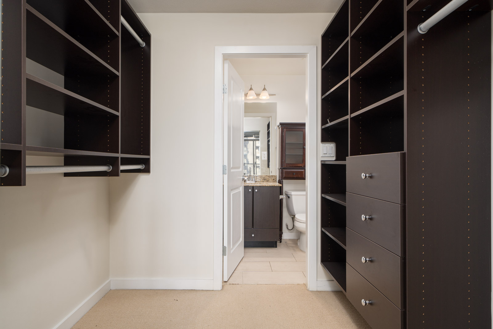 Walk-in closet with dark wood shelves and drawers, leading to a bathroom with a sink and mirror.