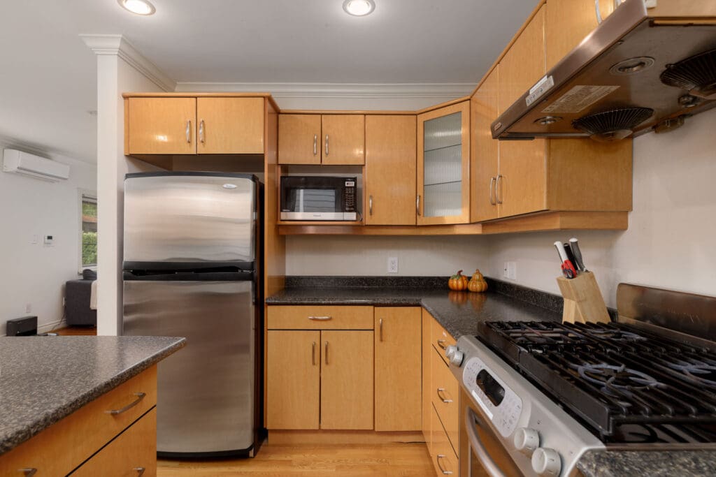 A modern kitchen with wooden cabinets, a stainless steel fridge, microwave, and a gas stove with a vent hood. A knife block is on the counter beside small decorative pumpkins.