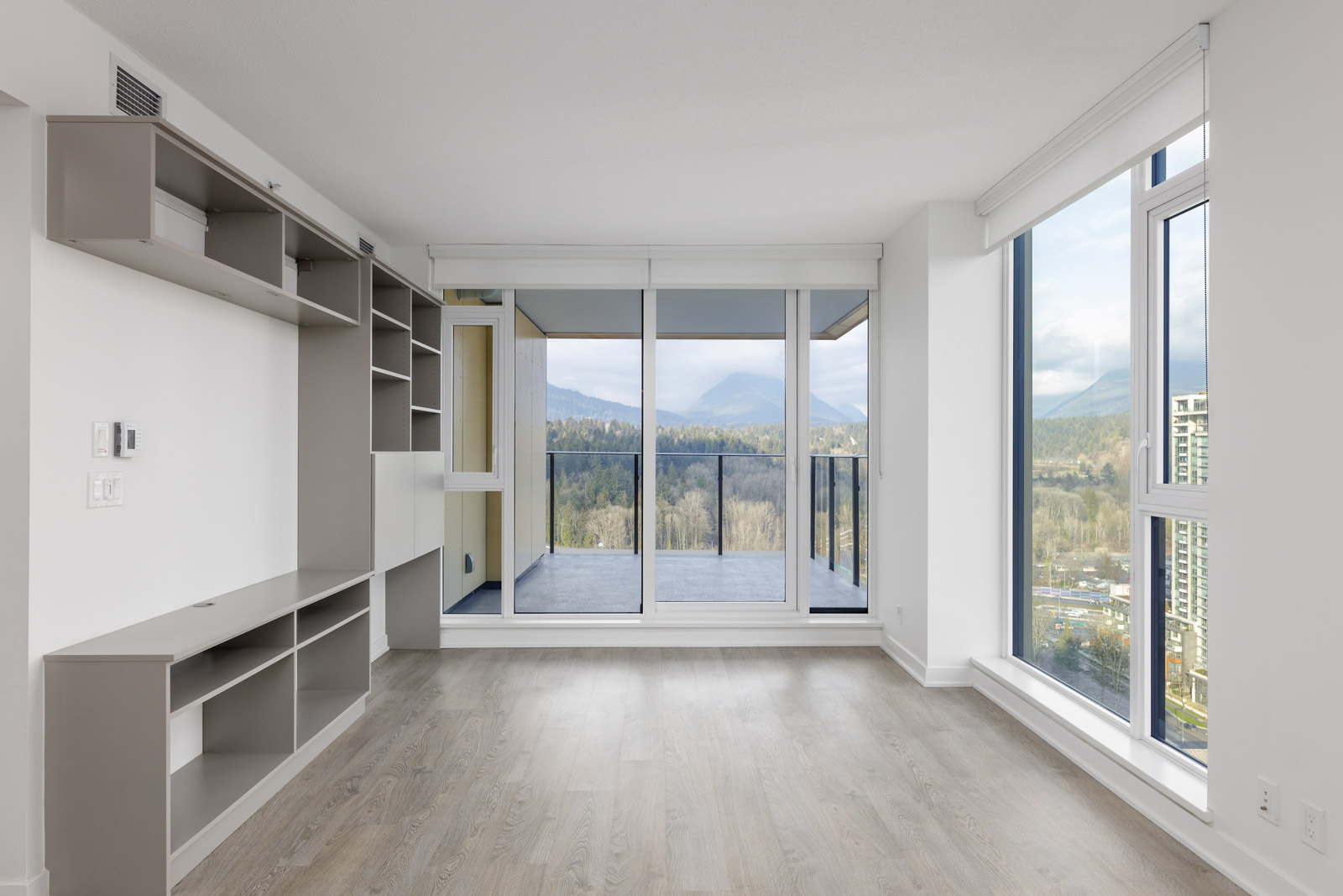 Empty modern apartment with large windows, built-in shelves, and a balcony offering a view of trees and mountains.
