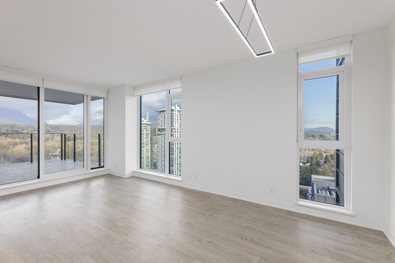 Empty apartment room with large windows and light wood flooring, offering a view of a cityscape and distant mountains. A balcony is visible on the left side.