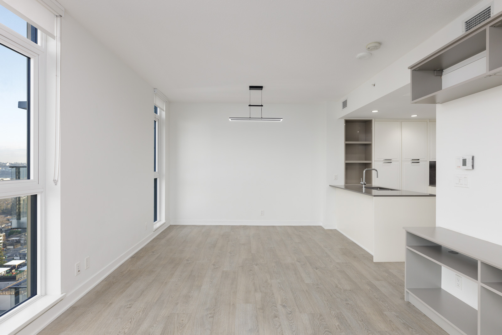 Empty modern apartment interior with light wood flooring, large windows, a kitchen area with white cabinets, and a ceiling light fixture.